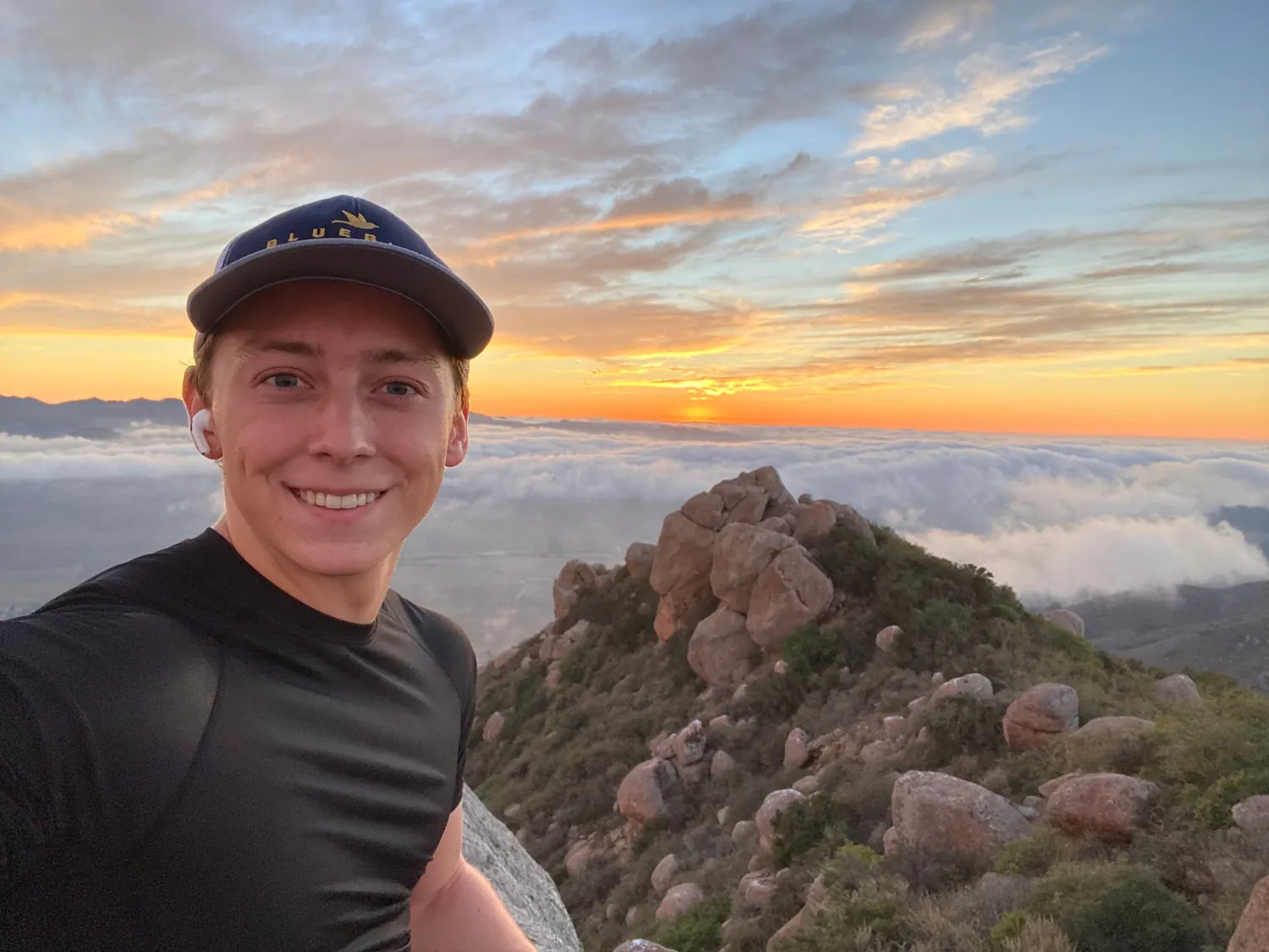 Sebastian smiling on a rocky peak above a sea of clouds at sunset